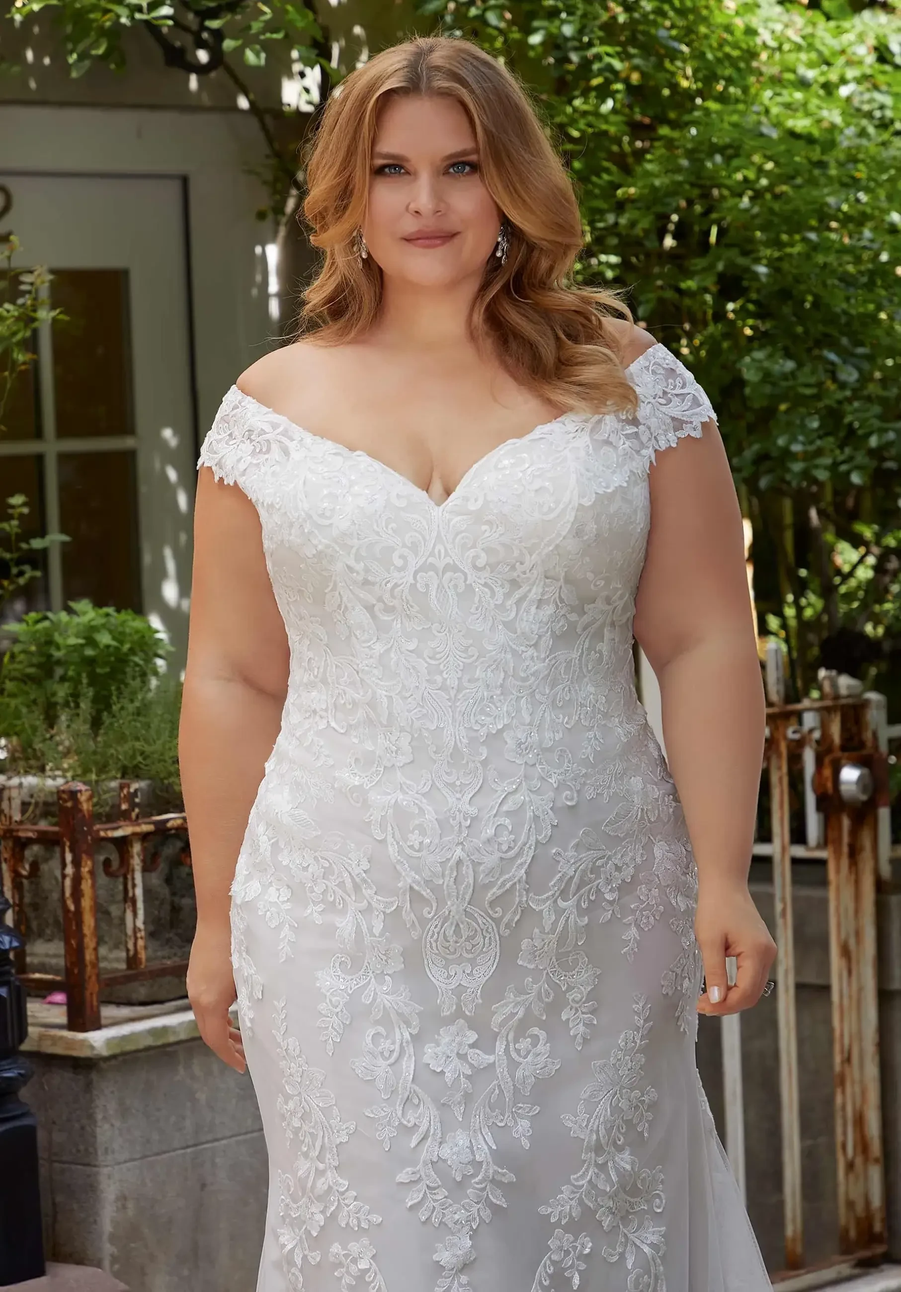 A woman in a detailed white wedding dress stands outdoors, smiling at the camera. The dress has an off-shoulder design and intricate lace patterns. She has wavy, shoulder-length hair and is surrounded by greenery.
