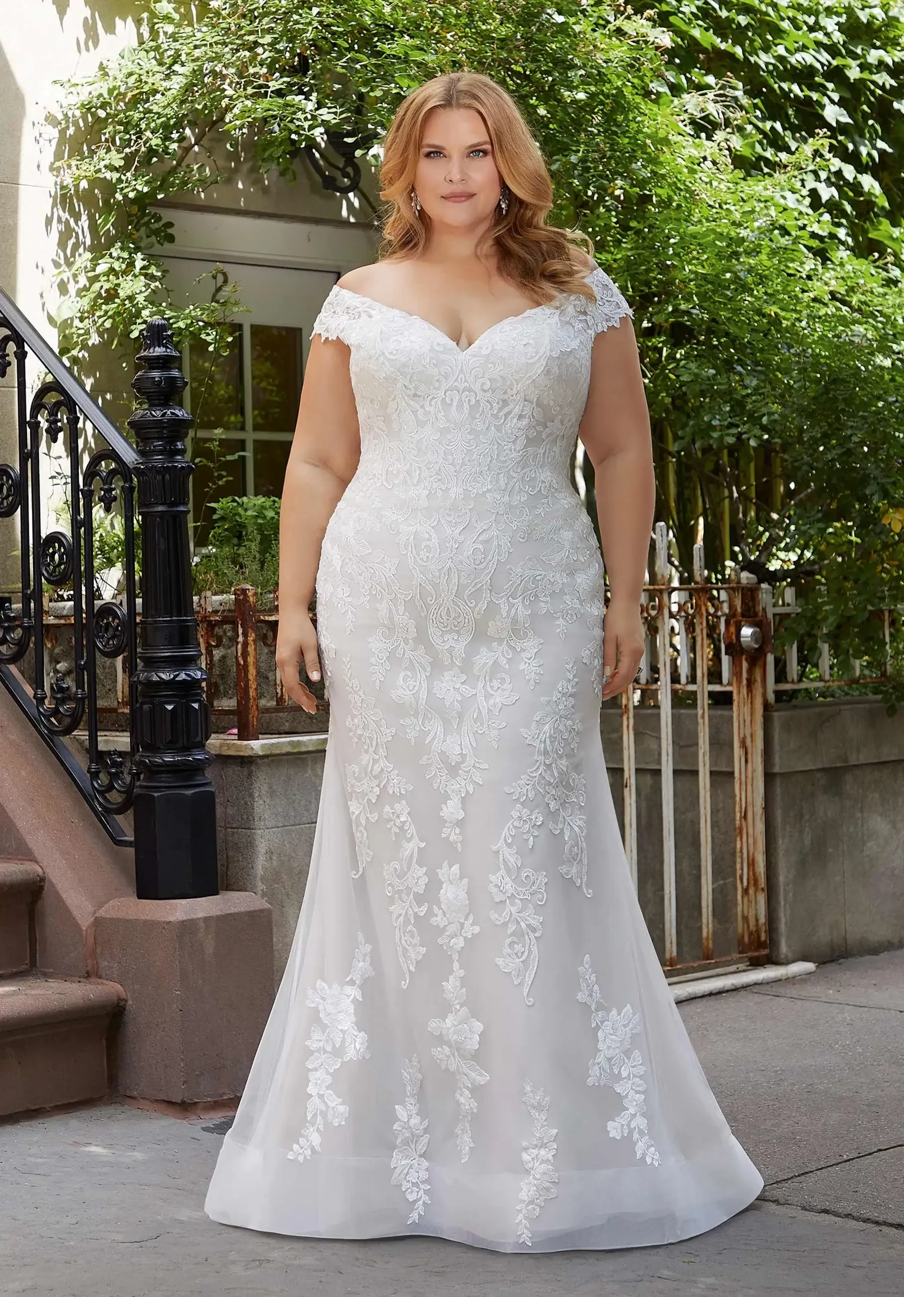 A woman in an elegant off-the-shoulder white wedding dress with intricate lace details, standing on steps outside a building surrounded by greenery.