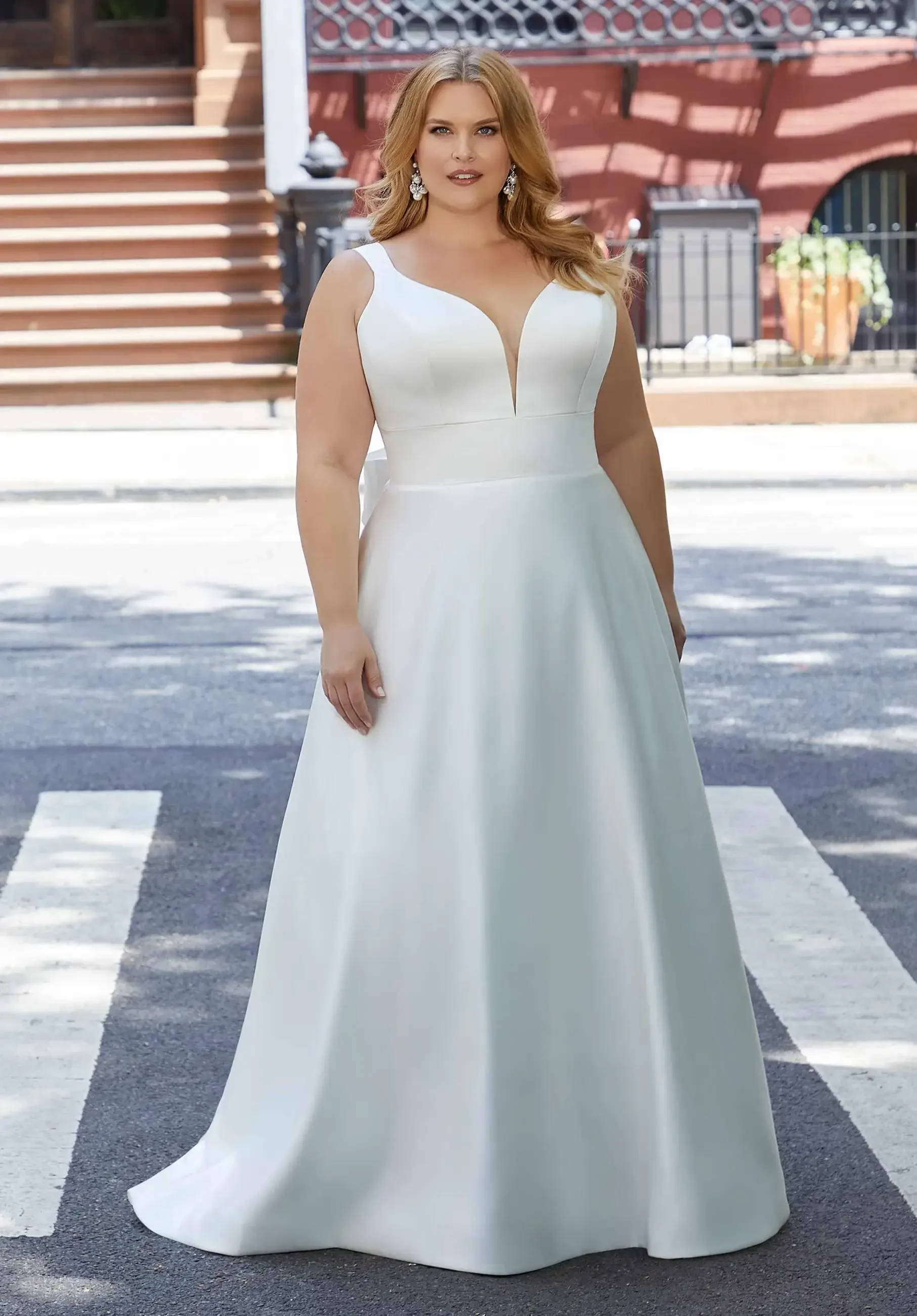 A woman wearing a white gown with a deep V-neck and wide straps, standing on a city street.