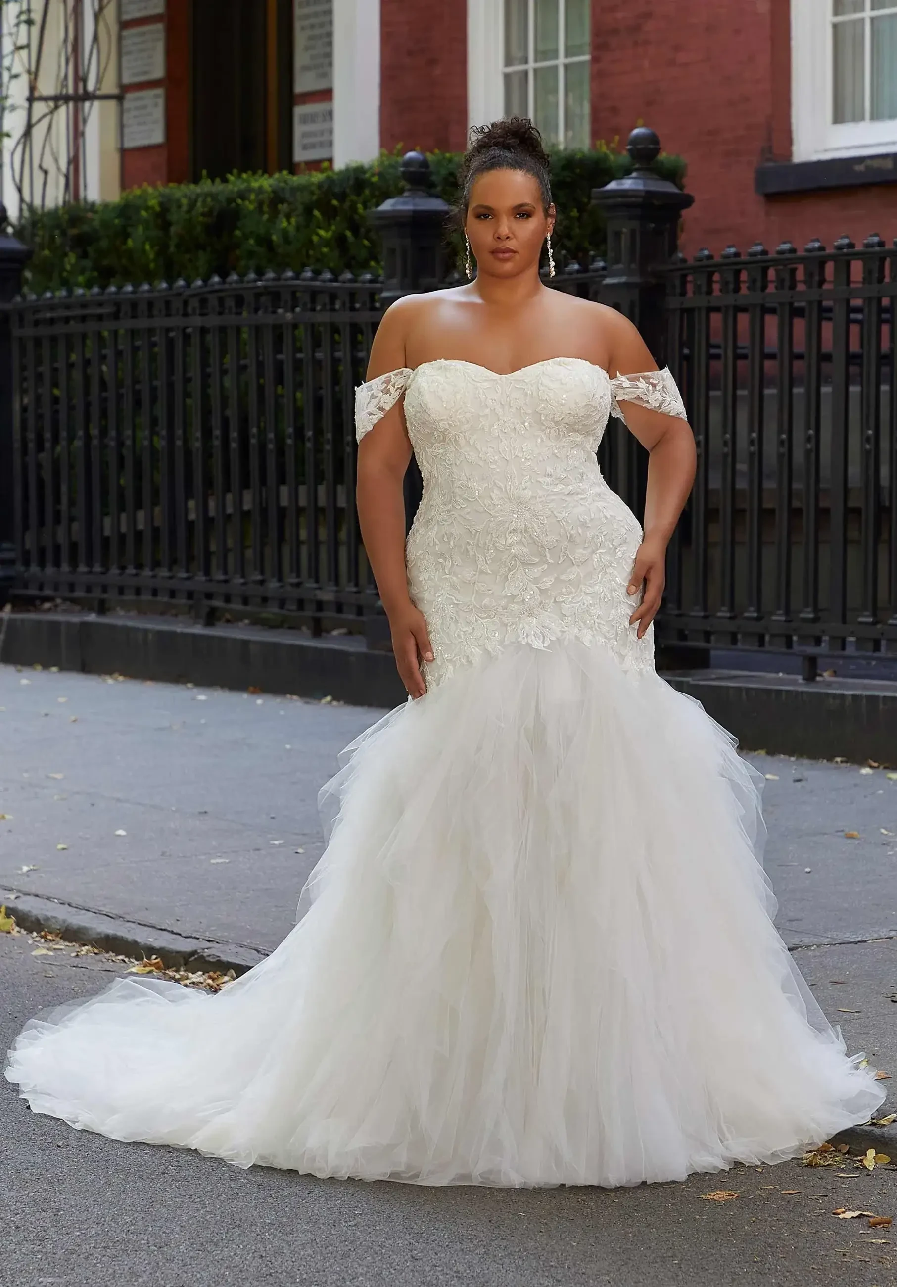 A woman wearing a strapless, off-the-shoulder wedding dress with lace details and a tulle skirt, standing on a city street.