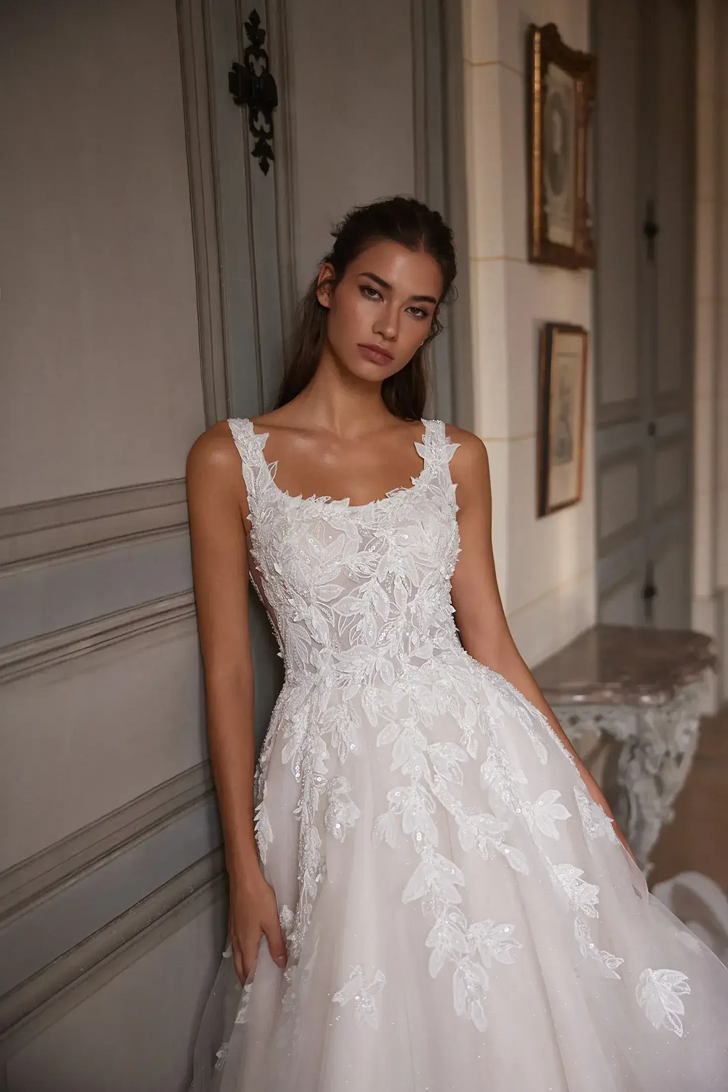 Bride in an elegant white wedding dress with floral lace detail stands in a vintage room, exuding a serene and romantic ambiance.