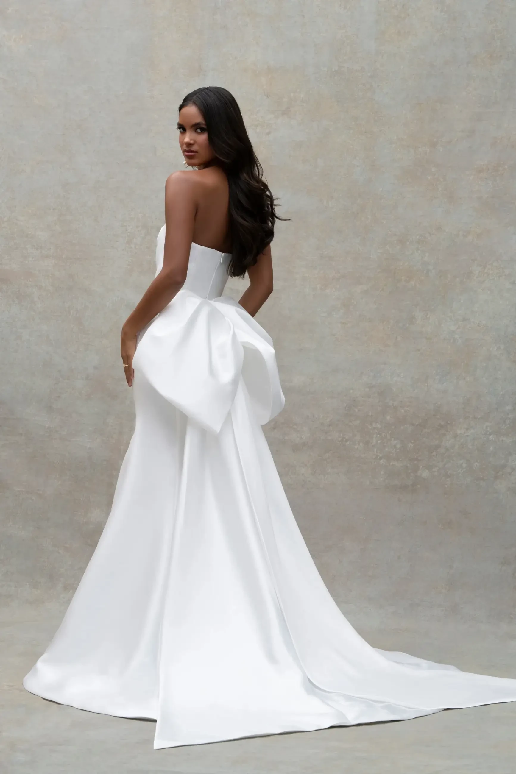 Model is wearing a sheath strapless bridal gown with a statement bow at the train