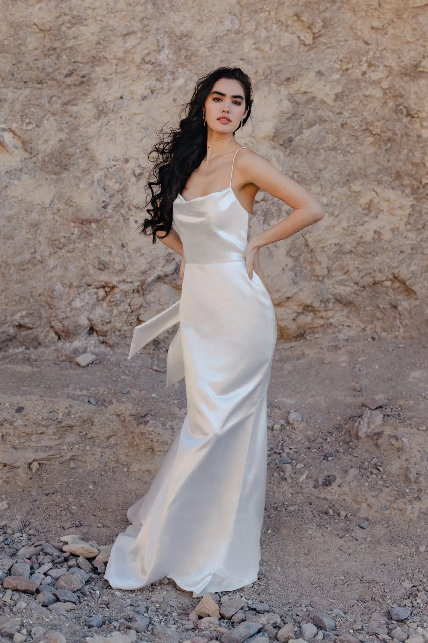 Model is wearing a spaghetti strap wedding dress with a bow at the waist and a sweep train