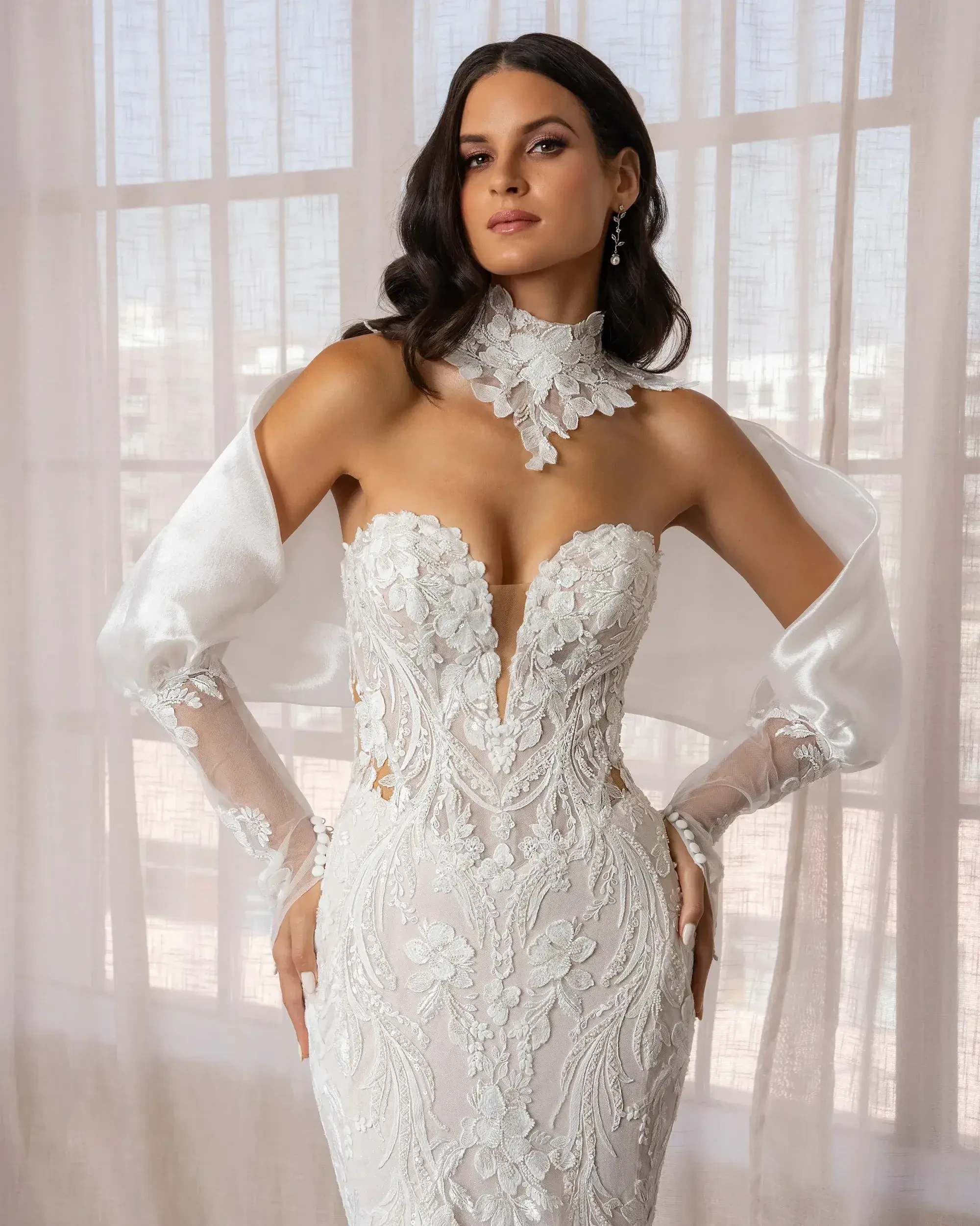 Woman in a white, floral lace gown stands confidently. The dress features a deep neckline, silk sleeves, and a lace choker. Soft light filters through sheer curtains.