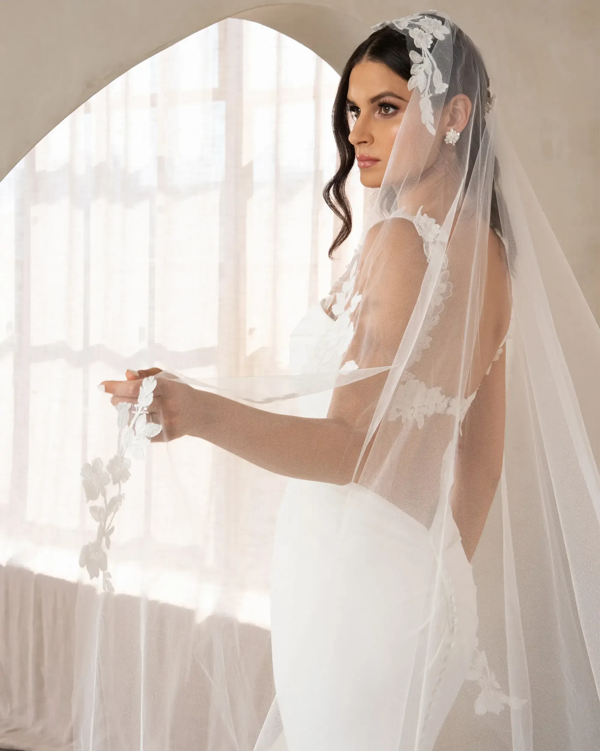 A bride wearing a fitted white gown and a decorative veil, standing in front of a large window with soft light filtering in.