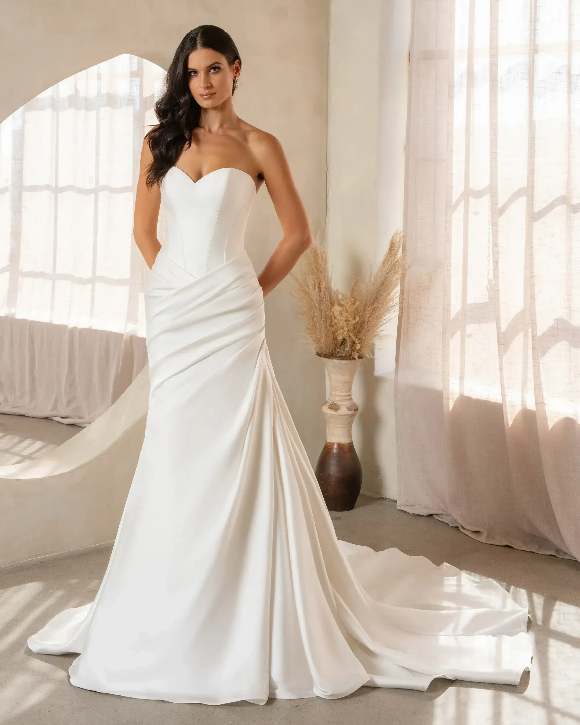 A model wearing a strapless white wedding dress with a fitted bodice and flowing skirt, standing in a well-lit room with natural decor.