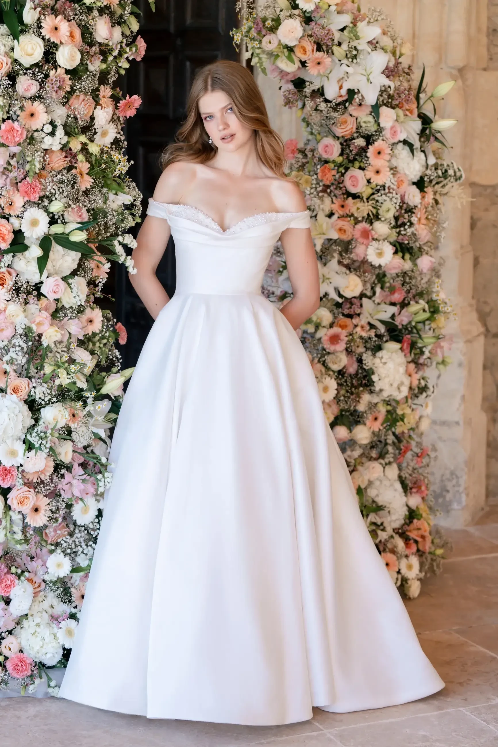 Model is wearing an off-shoulder Mikado ballgown with pearl and floral sequin embellishments along the sweetheart neckline.